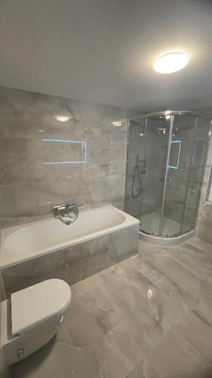 Wykończona łazienka z wanną, kabiną prysznicową z zaokrąglonymi drzwiami i podwieszaną toaletą, wyłożona płytkami imitującymi marmur w odcieniach szarości, oświetlona centralnym, okrągłym plafonem.