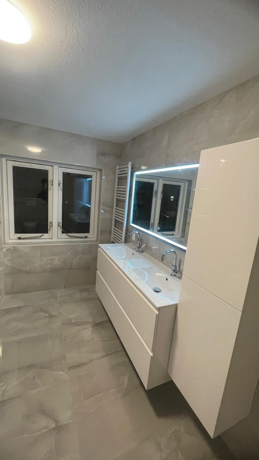 Nowoczesna łazienka z podwójną umywalką w białej szafce, podświetlanym lustrem, grzejnikiem drabinkowym i płytkami imitującymi marmur na ścianach i podłodze, widok z boku