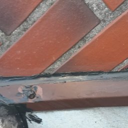 Złota rączka - Damian Krawczyk - Fragment muru z czerwonej cegły ułożonej we wzór jodełki, uszczelniony czarnym silikonem, widoczne ślady rdzy i zabrudzenia.