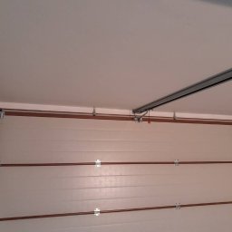 Złota rączka - Damian Krawczyk - Wnętrze garażu z widoczną częściowo otwartą segmentową bramą garażową, szynami prowadzącymi i mocowaniami do sufitu.