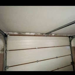 Złota rączka - Damian Krawczyk - Biała segmentowa brama garażowa z widocznymi śladami wilgoci i pleśni na górnej krawędzi oraz na ścianie obok, z metalowymi prowadnicami i fragmentem sufitu.