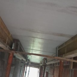 Złota rączka - Damian Krawczyk - Betonowy strop w trakcie budowy, podparty metalowymi stemplami i drewnianymi belkami, widoczne ślady wilgoci na suficie, surowy stan wykończenia.