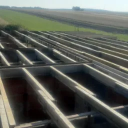 Wiązanie drewnianych belek stropowych na murze z czerwonej cegły, widok z góry na tle pól uprawnych.