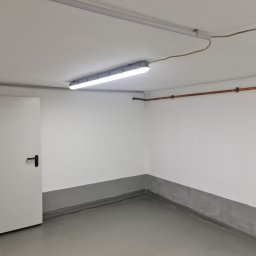 Złota rączka - Damian Krawczyk - Pomalowane na biało pomieszczenie z szarą lamperią, widoczna instalacja elektryczna pod sufitem i rura miedziana na ścianie, oświetlenie jarzeniowe i białe drzwi z czarną klamką.