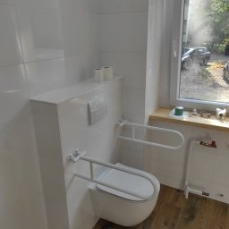 Złota rączka - Damian Krawczyk - Biała, podwieszana toaleta z uchwytami dla niepełnosprawnych w wykafelkowanej łazience z drewnopodobną podłogą, widok na okno z zielenią za szybą.