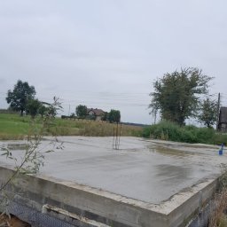 Złota rączka - Damian Krawczyk - Wylany fundament pod budynek jednorodzinny, z widoczną hydroizolacją i wystającymi prętami zbrojeniowymi, w tle zielony krajobraz i dom.