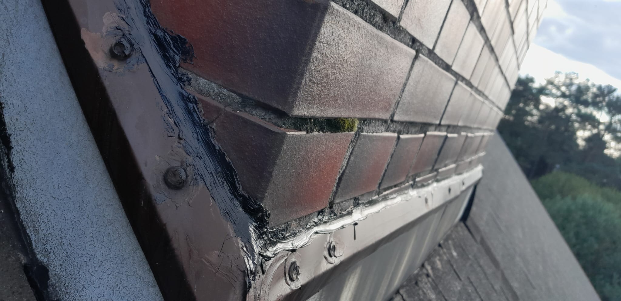 Szczegółowe ujęcie odnowionego komina z cegieł, z widocznymi śladami świeżo nałożonego uszczelniacza i metalową obróbką blacharską mocowaną śrubami, z mchem w spoinach i fragmentem dachu w tle.