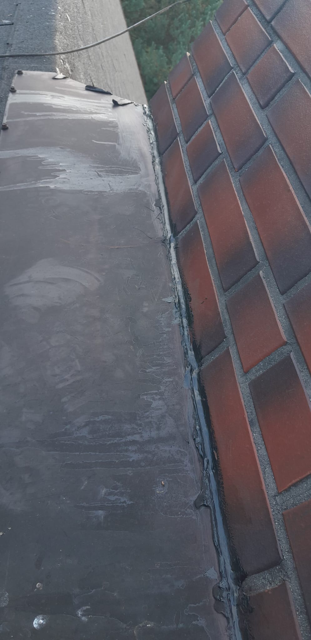 Połączenie papy bitumicznej z murem z czerwonej cegły, uszczelnione czarną masą bitumiczną, widoczne ślady wody.