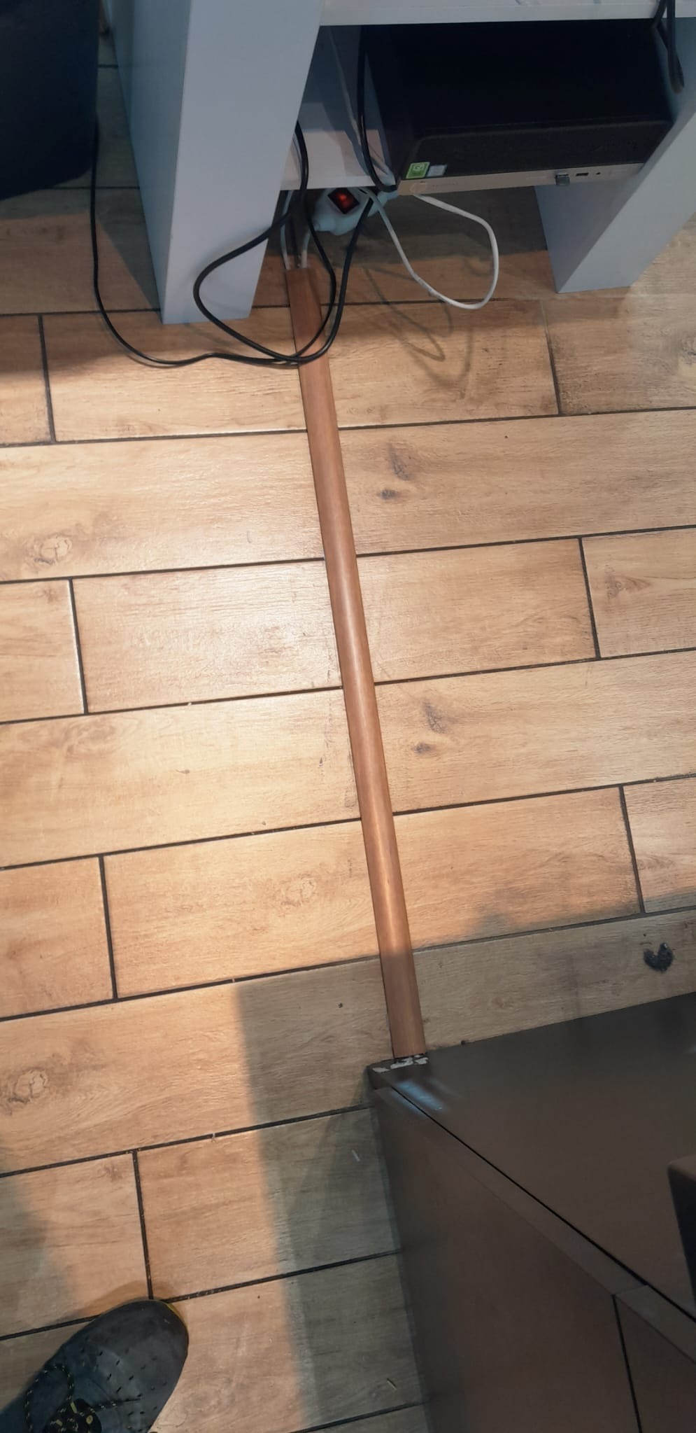 Brązowa listwa przypodłogowa z PCV na tle płytek imitujących drewno, widoczne kable i fragment nogi mebla oraz buta.
