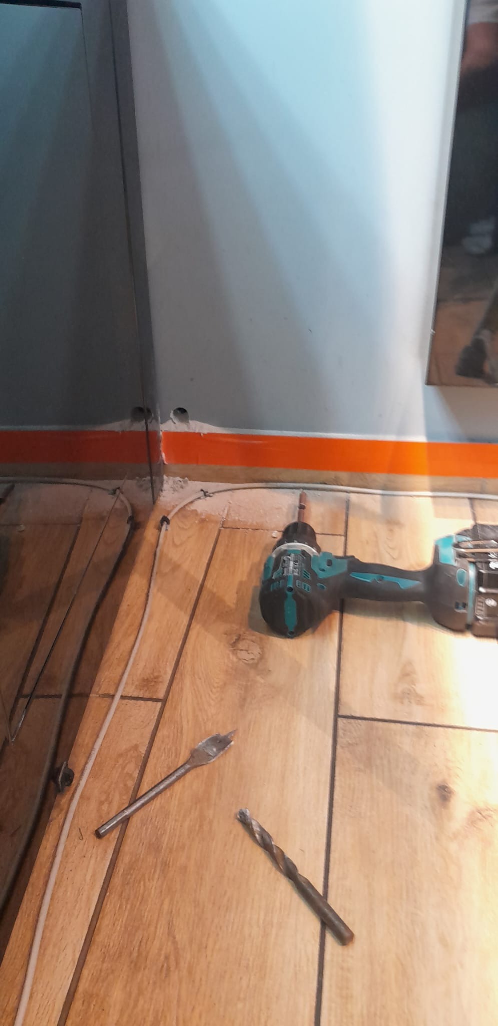 Wiertarka akumulatorowa, wiertła i rurki grzewcze na drewnianej podłodze, widoczne otwory w ścianie i listwa przypodłogowa.