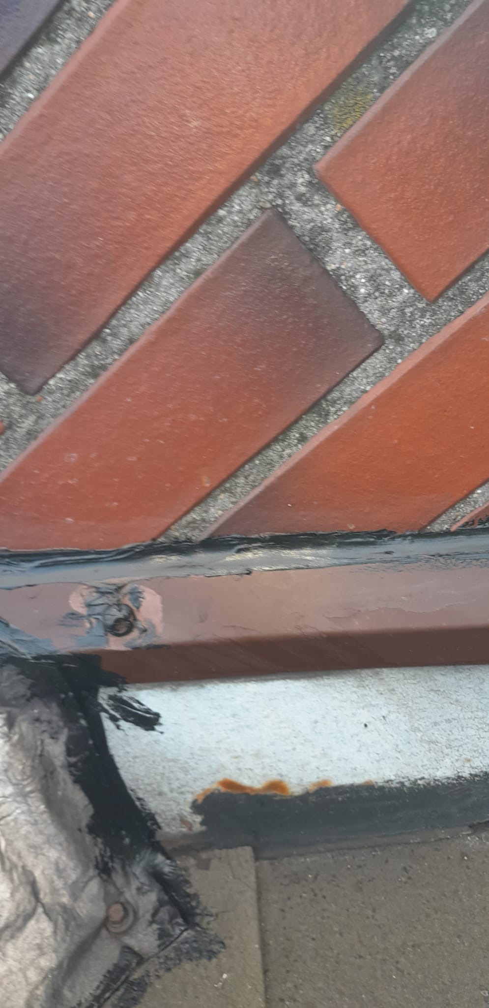 Fragment muru z czerwonej cegły ułożonej we wzór jodełki, uszczelniony czarnym silikonem, widoczne ślady rdzy i zabrudzenia.