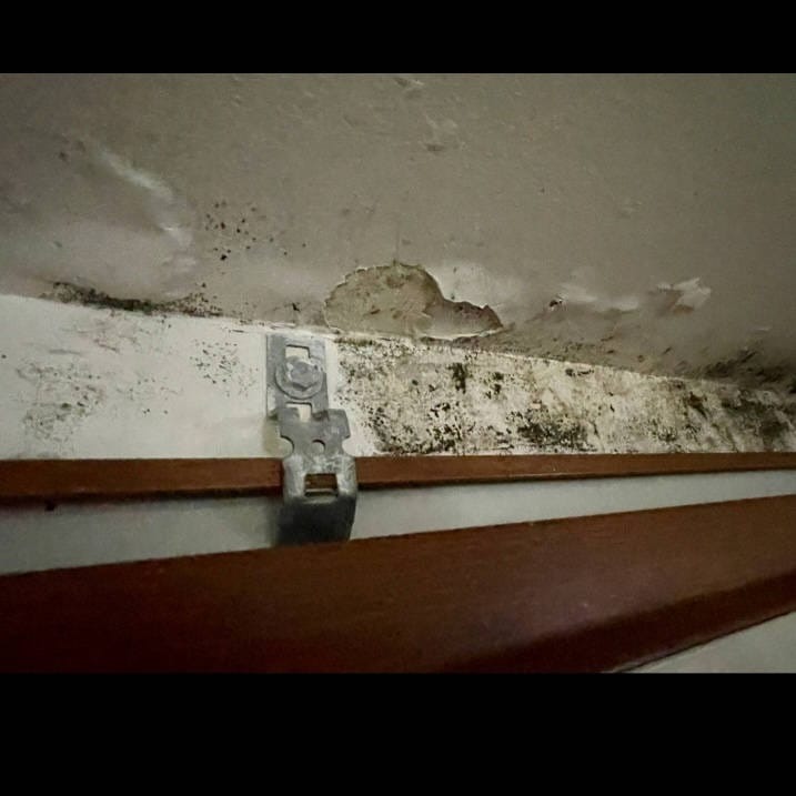 Zacieki i pleśń na suficie oraz ścianie, widoczny metalowy uchwyt mocujący.