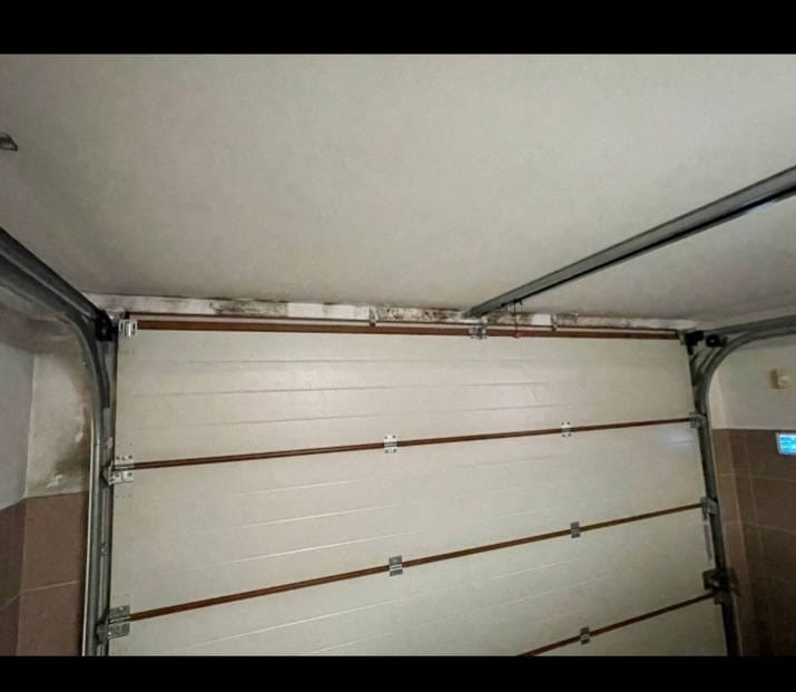 Biała segmentowa brama garażowa z widocznymi śladami wilgoci i pleśni na górnej krawędzi oraz na ścianie obok, z metalowymi prowadnicami i fragmentem sufitu.