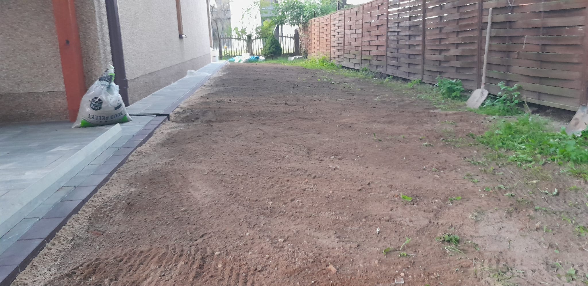 Przygotowany teren pod trawnik z widoczną kostką brukową, workiem z pelletem i łopatą opartą o drewniany płot.