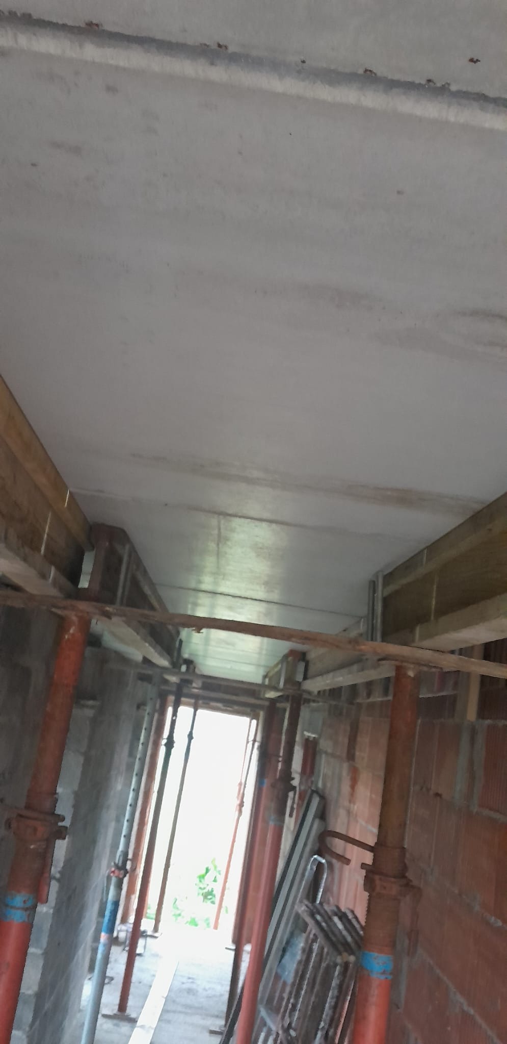 Betonowy strop w trakcie budowy, podparty metalowymi stemplami i drewnianymi belkami, widoczne ślady wilgoci na suficie, surowy stan wykończenia.