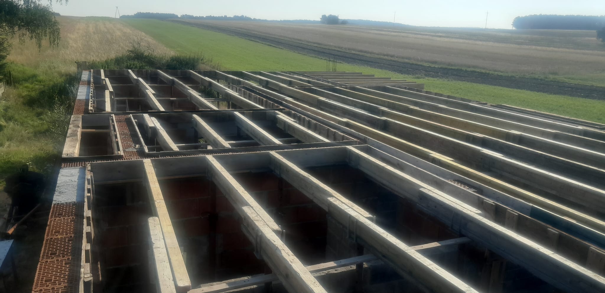 Wiązanie drewnianych belek stropowych na murze z czerwonej cegły, widok z góry na tle pól uprawnych.