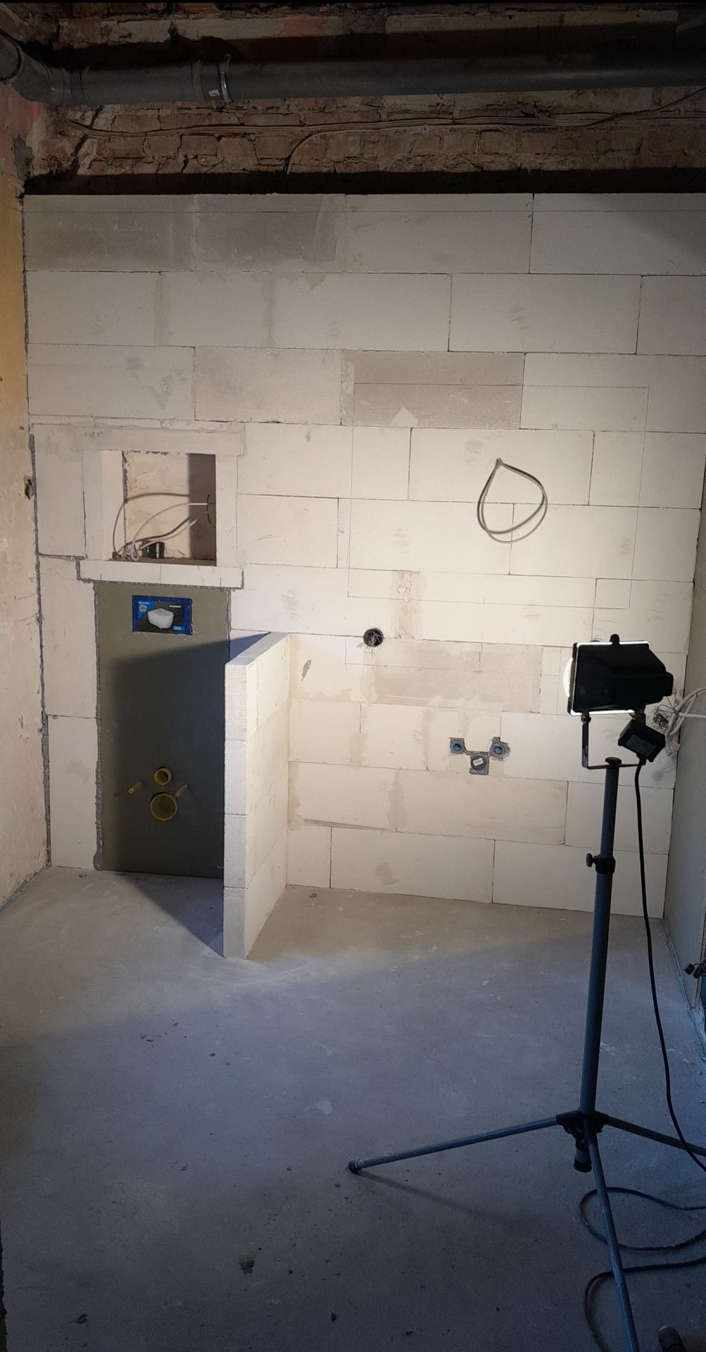 Surowy stan łazienki w trakcie budowy, ściany z białych bloczków, instalacja podtynkowego WC, widoczny reflektor na statywie.