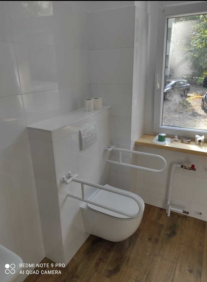 Biała, podwieszana toaleta z uchwytami dla niepełnosprawnych w wykafelkowanej łazience z drewnopodobną podłogą, widok na okno z zielenią za szybą.