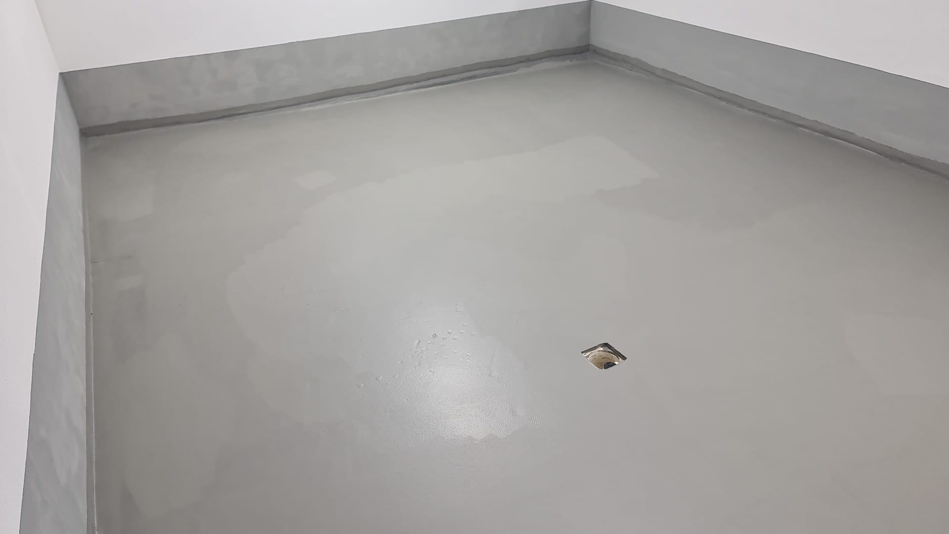 Świeżo wylany, szary beton samopoziomujący na podłodze w pomieszczeniu, widoczny otwór kanalizacyjny w narożniku.