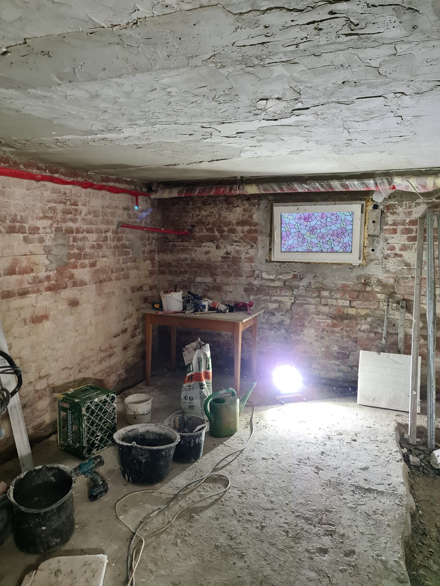 Piwnica w trakcie remontu: widoczne surowe ściany z cegły, niedokończony sufit, narzędzia i materiały budowlane rozłożone na podłodze i stole, okno z kolorową folią.