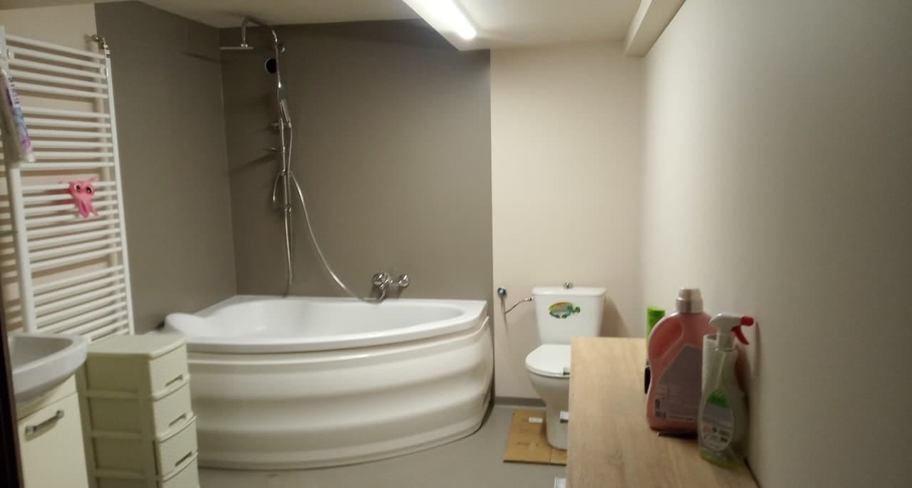 Wykończona łazienka z narożną wanną, grzejnikiem drabinkowym z zawieszoną różową zabawką, toaletą i szafką z umywalką, utrzymana w szaro-białej kolorystyce.