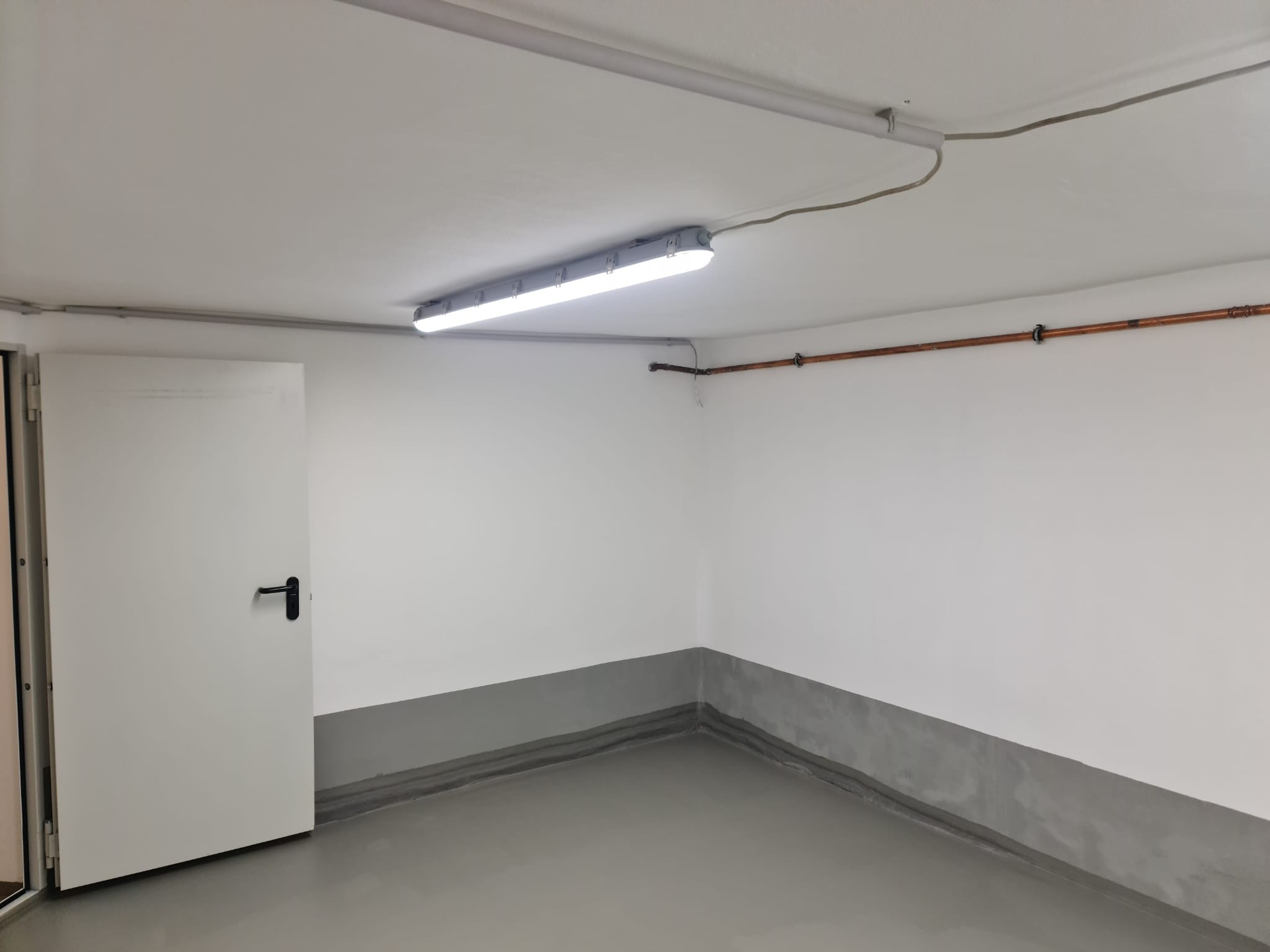 Pomalowane na biało wnętrze garażu z szarą lamperią, widoczna lampa jarzeniowa na suficie, rury miedziane przy ścianie i białe drzwi z czarną klamką.