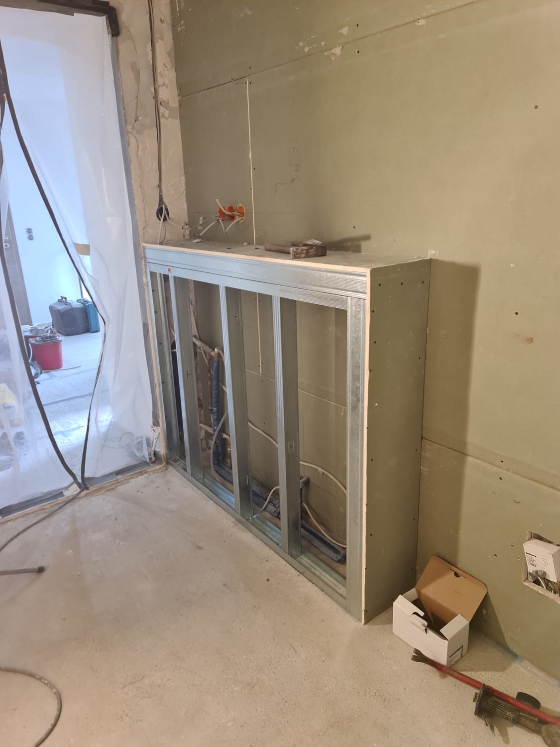 Wnętrze w trakcie prac wykończeniowych: metalowy stelaż zabudowy z widocznymi instalacjami, częściowo obłożony płytami gipsowo-kartonowymi, młotek na górnej krawędzi, surowe ściany i podłoga...