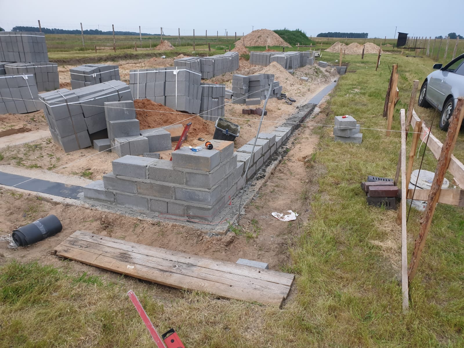 Wznoszenie fundamentów budynku z bloczków betonowych, widoczne zapasy materiału, poziomica, oraz częściowo wykonana ściana fundamentowa na tle krajobrazu polnego.