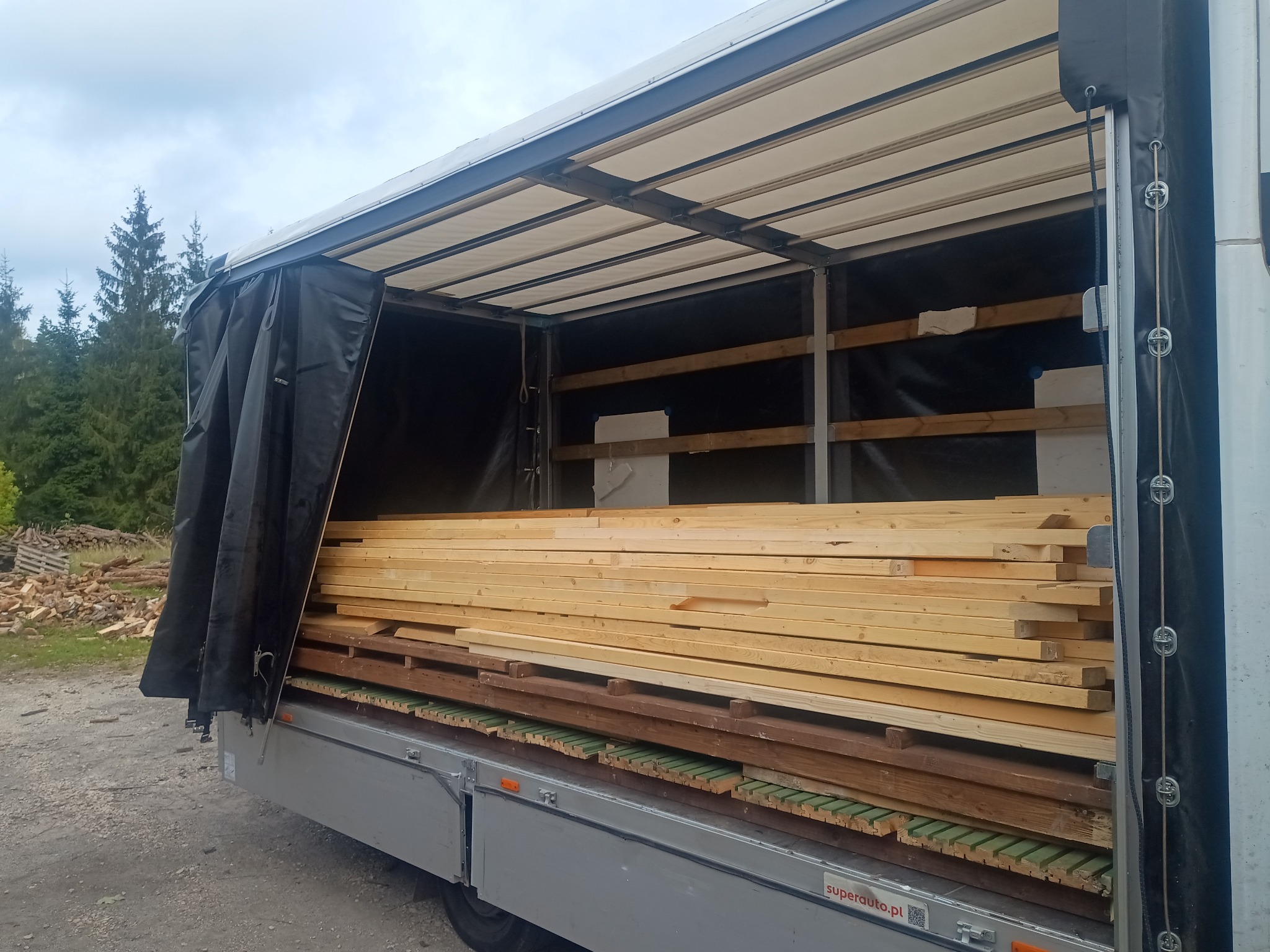 Częściowo otwarty bus z załadowanym drewnem budowlanym, widoczne kantówki i deski na tle lasu.