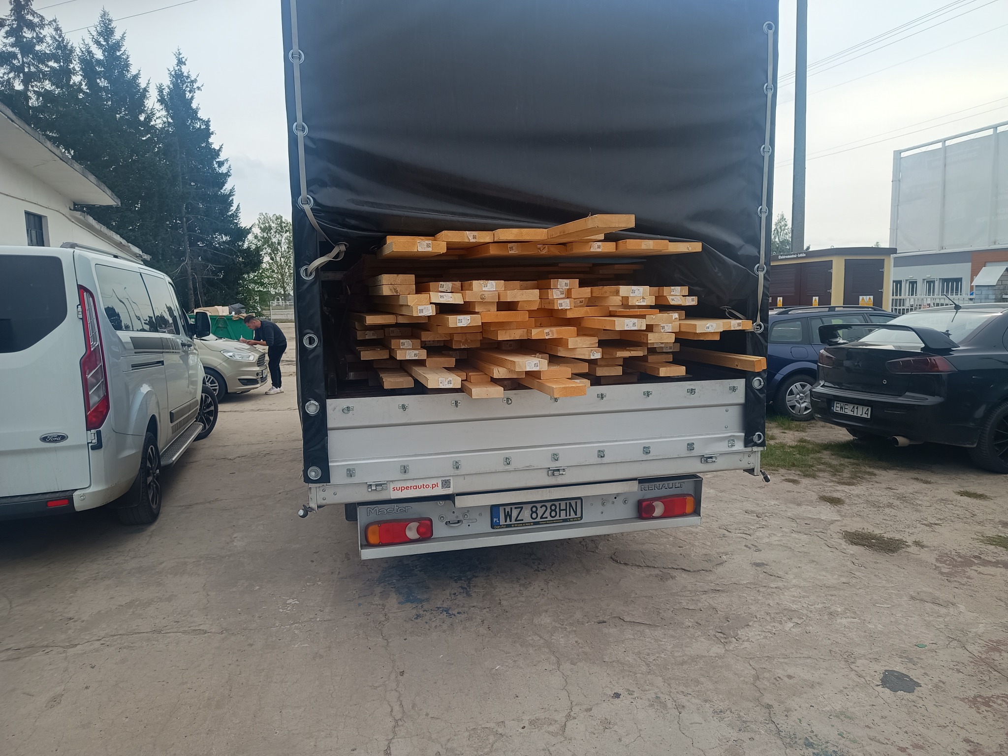 Załadunek drewna do busa z czarną plandeką, widoczny numer rejestracyjny WZ 828HN, obok biały Ford Transit i inne zaparkowane auta.