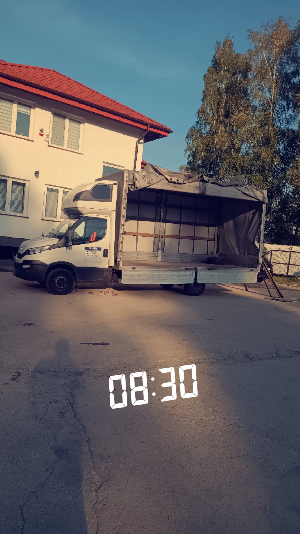 Biały samochód dostawczy z otwartą przestrzenią ładunkową zaparkowany na ulicy przed budynkiem z czerwonym dachem, widoczne oznaczenia firmy na kabinie.