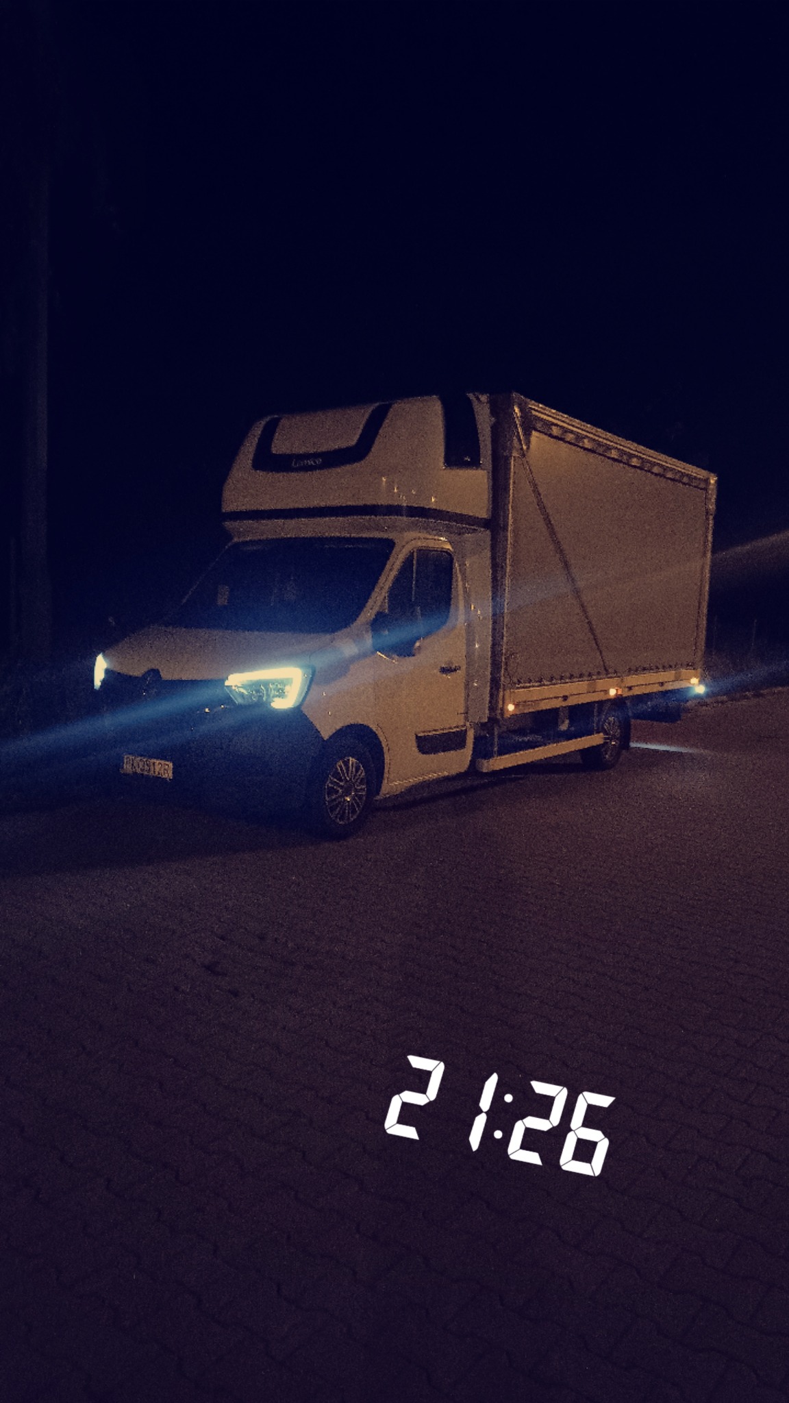 Biały bus dostawczy Renault Master z wysoką zabudową kontenerową, sfotografowany nocą na oświetlonej ulicy, z widocznym numerem rejestracyjnym i cyfrowym zegarem 21:26 w dolnej części kadru.