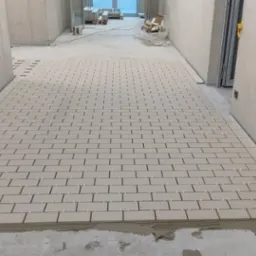 Układanie jasnych, prostokątnych płytek na podłodze w korytarzu, widoczne krzyżyki dystansowe i świeży klej.