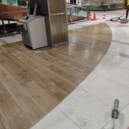 UNI-FLOOR Dominik Jaworowski - Montaż drewnopodobnych płytek podłogowych w przestrzeni handlowej, widoczne narzędzia i materiały budowlane, pracownik w tle.