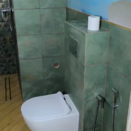 Nowoczesna toaleta podwieszana z bidetką, obudowana zielonymi płytkami. Na g&oacute;rze widoczna rolka papieru toaletowego. Podłoga wyłożona drewnopodobnymi panelami w jodełkę.