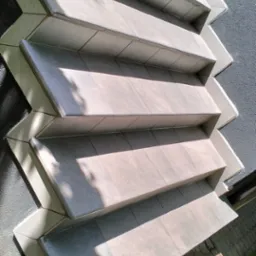 Kaskadowe schody zewnętrzne wyłożone szarą płytką, z widocznym fragmentem elewacji budynku o szarej strukturze i kostki brukowej. Ujęcie z dołu, w słoneczny dzień.