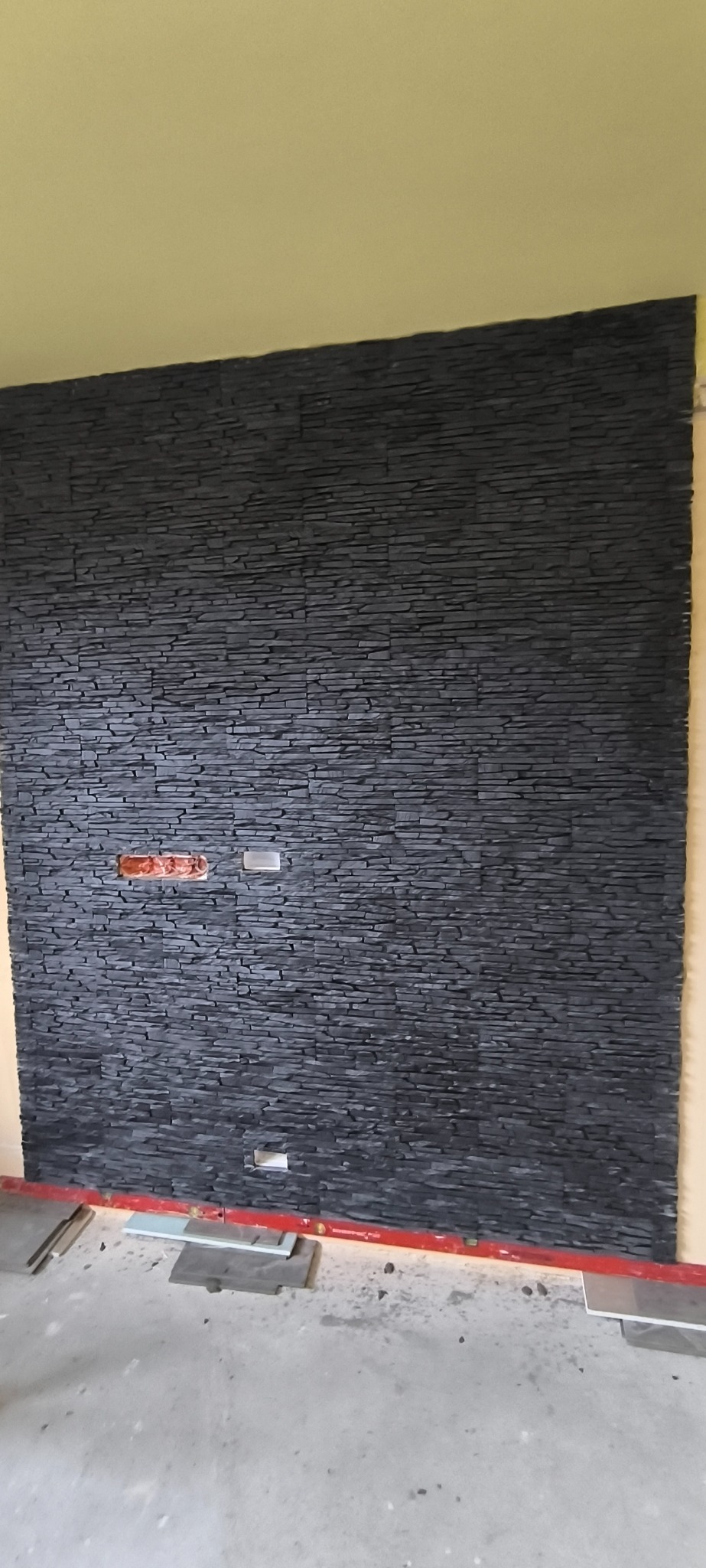 Ściana wykończona czarnym kamieniem dekoracyjnym o nieregularnej fakturze, z widocznymi puszkami elektrycznymi i poziomnicą na podłodze. Żółta ściana w tle.
