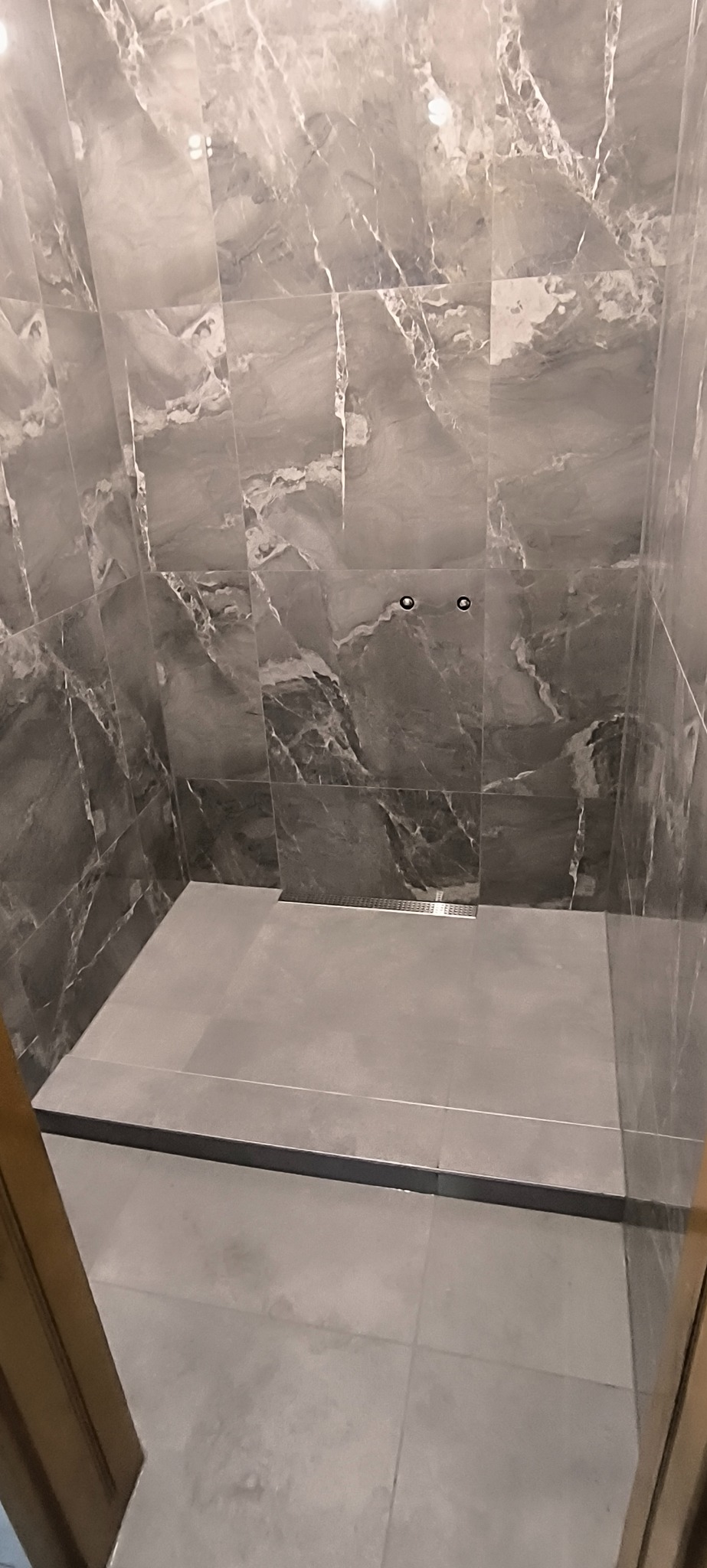 Kabina prysznicowa z szarymi płytkami imitującymi marmur, z odpływem liniowym. Widoczne dwa punkty mocowania baterii. Minimalistyczny design.