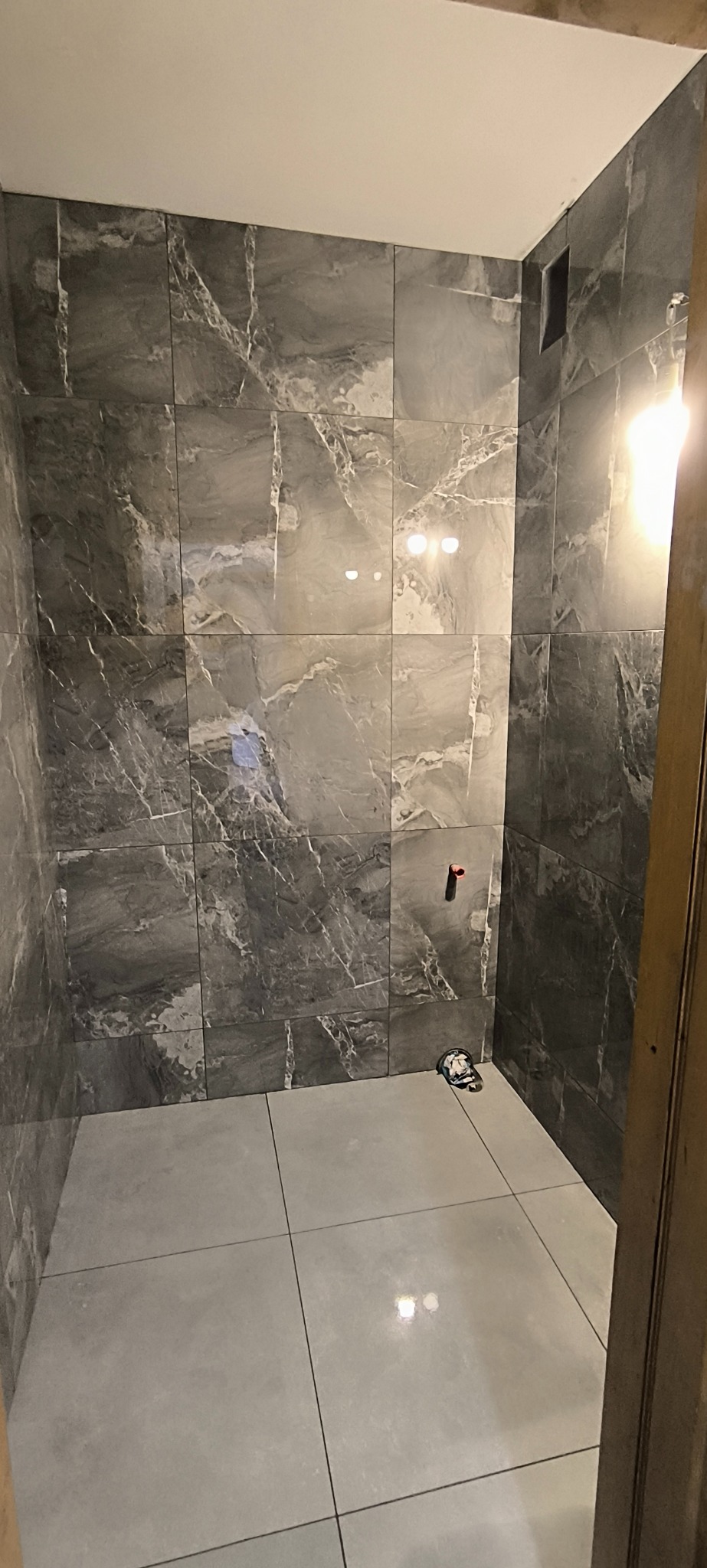 Wykończona kabina prysznicowa z szarymi płytkami imitującymi marmur na ścianach i jasnymi płytkami na podłodze. Widoczny fragment instalacji wodnej.