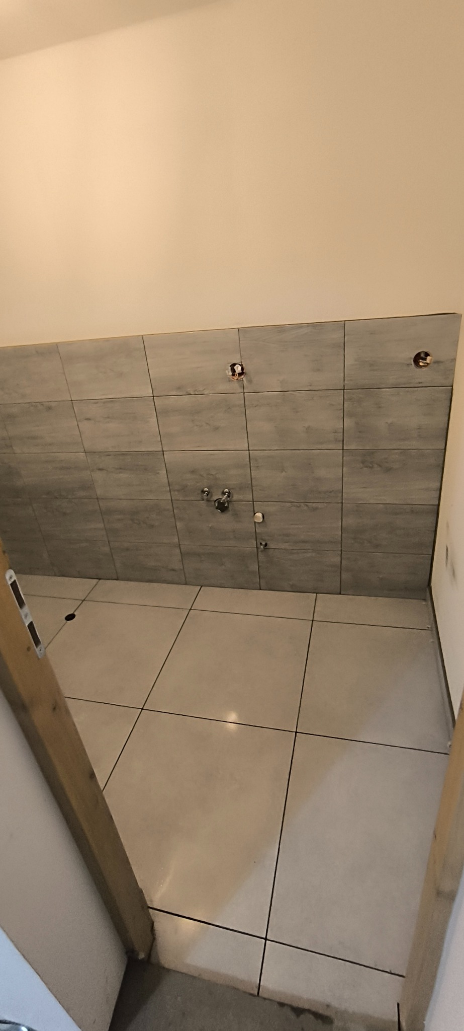 Wykończona ściana i podłoga w łazience z szarymi płytkami imitującymi drewno na ścianie i jasnymi płytkami na podłodze. Widoczne wyprowadzenia instalacji.