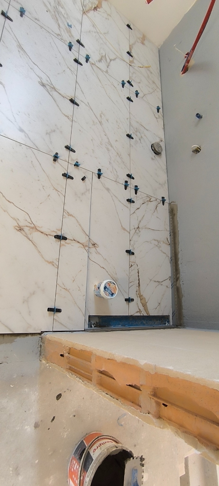 Układanie płytek imitujących marmur na ścianie z użyciem klipsów poziomujących. Widoczny odpływ liniowy i wiaderko z klejem. Prace wykończeniowe w łazience.