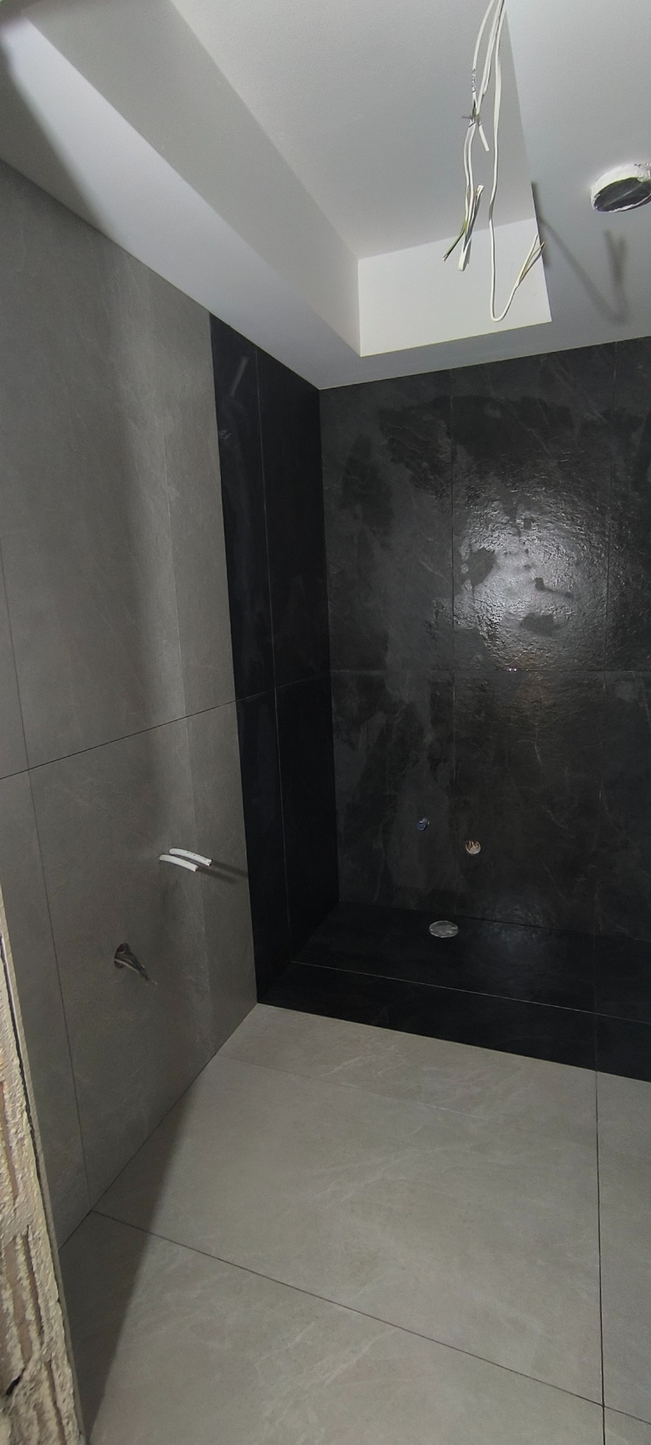 Nowoczesna kabina prysznicowa z szarymi i czarnymi płytkami, widoczne elementy instalacji. Minimalistyczny design, surowy styl wykończenia łazienki.