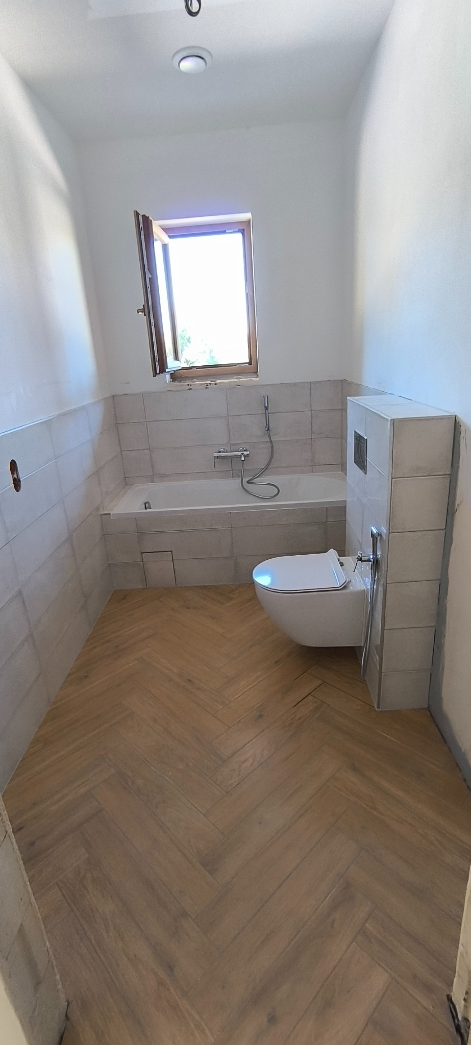 Jasna łazienka po remoncie: drewnopodobna podłoga w jodełkę, biała wanna i toaleta pod oknem. Minimalistyczny design z elementami wykończeniowymi.