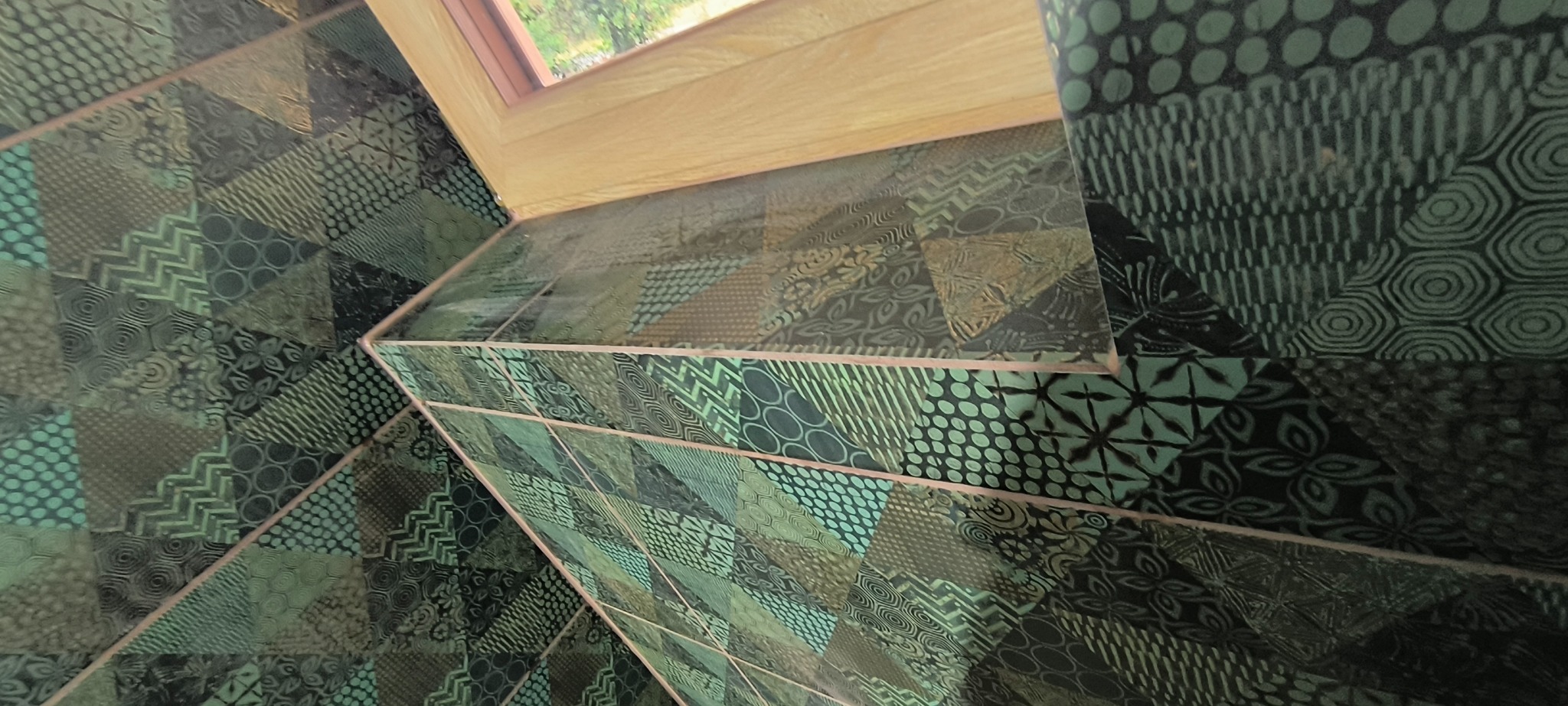 Ukończona łazienka z oryginalnymi, wzorzystymi, zielono-czarnymi płytkami ceramicznymi ułożonymi pod skosem, widok na okno dachowe.