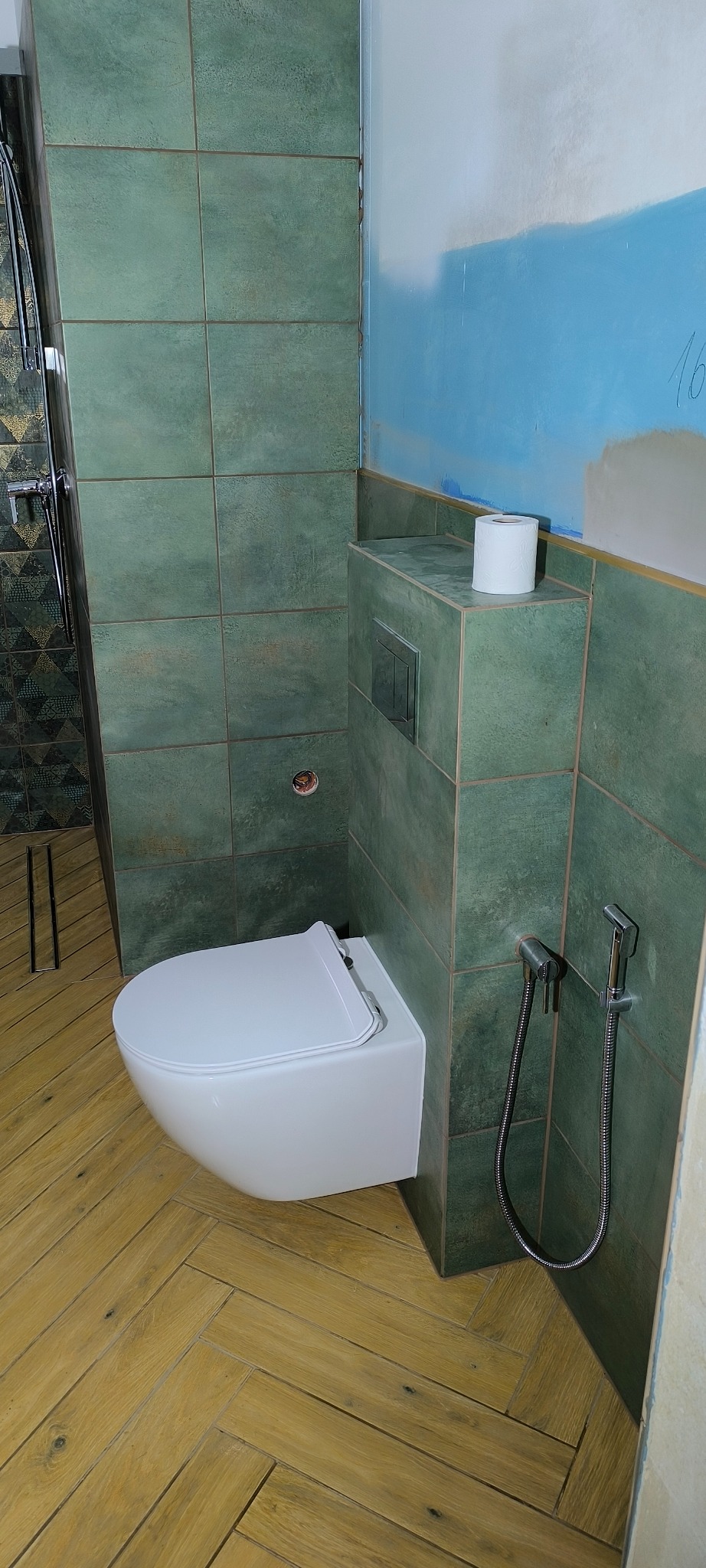 Nowoczesna toaleta podwieszana z bidetką, obudowana zielonymi płytkami. Na górze widoczna rolka papieru toaletowego. Podłoga wyłożona drewnopodobnymi panelami w jodełkę.