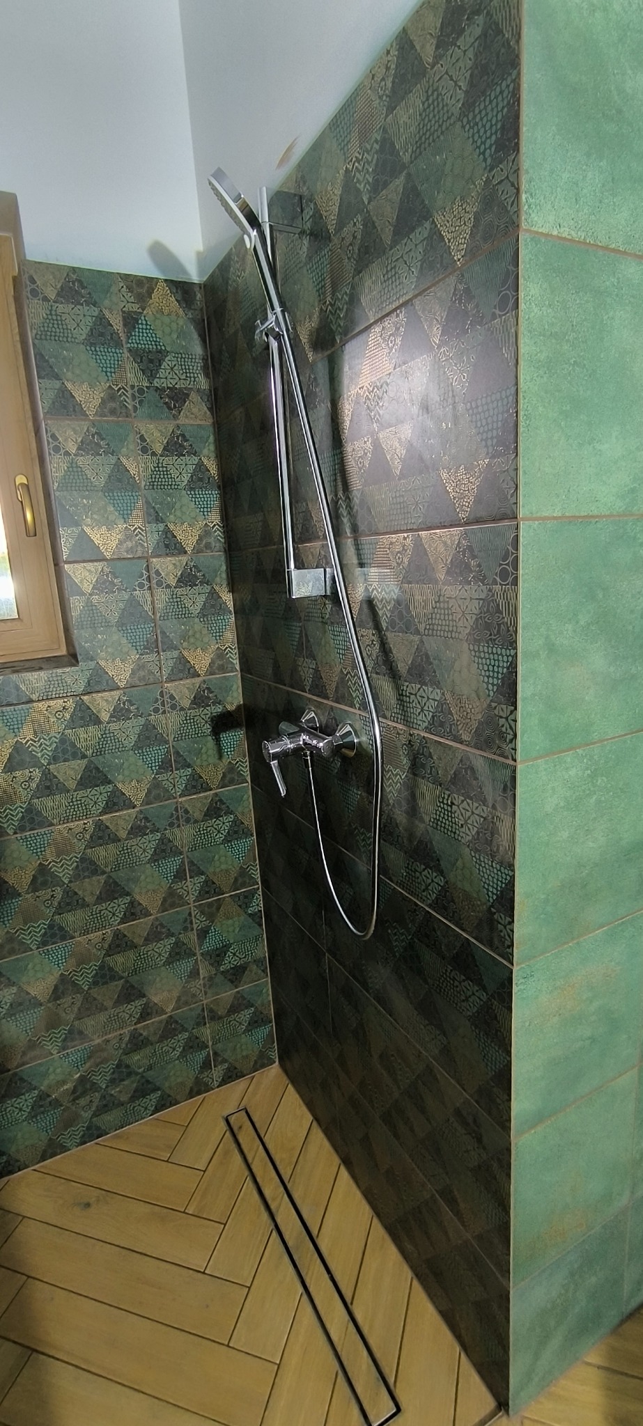 Kabina prysznicowa z zielono-złotymi płytkami o wzorze geometrycznym, chromowana armatura i odpływ liniowy. Podłoga wyłożona płytkami imitującymi drewno.