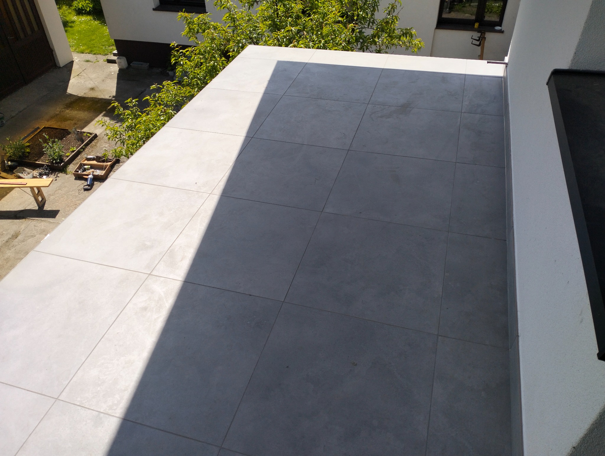 Wykończony taras z szarych, kwadratowych płytek w Dębnie, widziany z góry. Częściowo oświetlony słońcem, z widocznymi narzędziami i zielenią w tle.