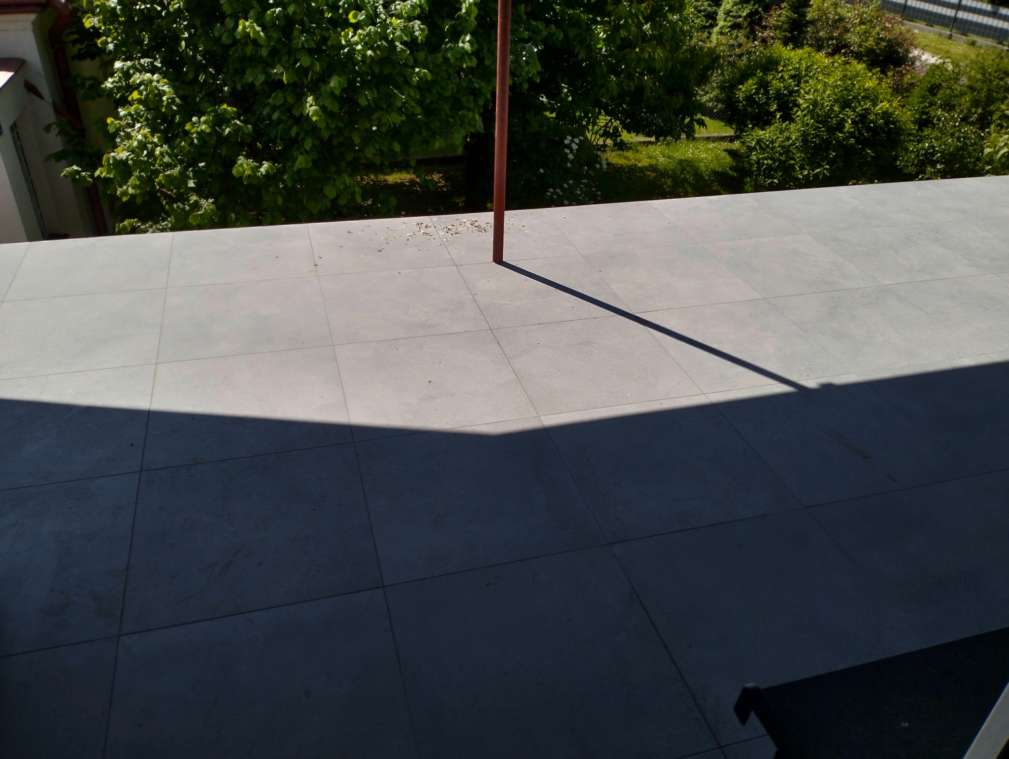 Szare płytki na balkonie w słoneczny dzień, cień słupa przecina kafelki. Widok z góry na świeżo wyłożoną podłogę balkonu z widocznym tłem zieleni.