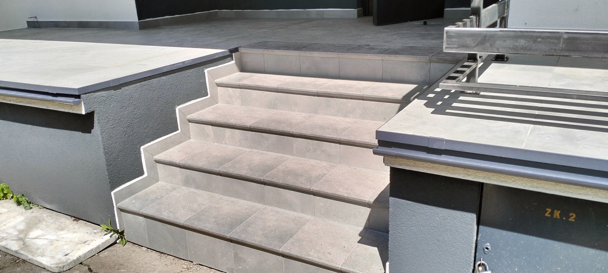 Wykończone płytkami schody zewnętrzne z szarymi fugami, prowadzące do wejścia. Widoczne detale wykończenia i fragment metalowej konstrukcji. Jasne, naturalne oświetlenie.