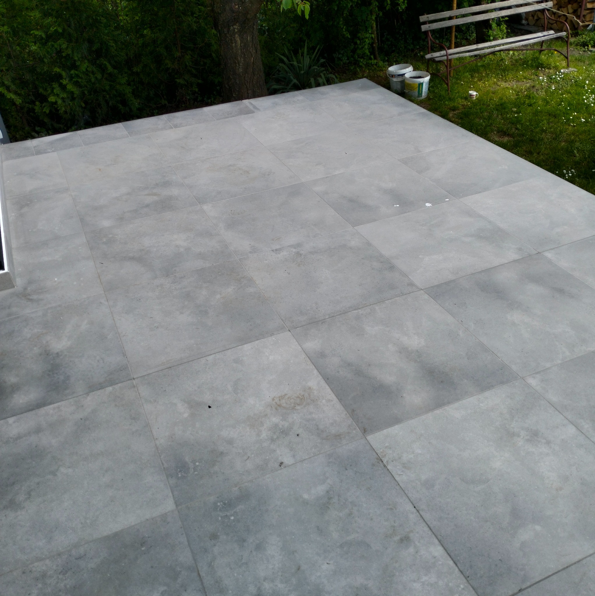Ułożona, szara, kwadratowa podłoga z płytek na zewnątrz, z widocznym fragmentem trawnika, drzewa i ławki w tle. Widoczne fugi między płytkami.